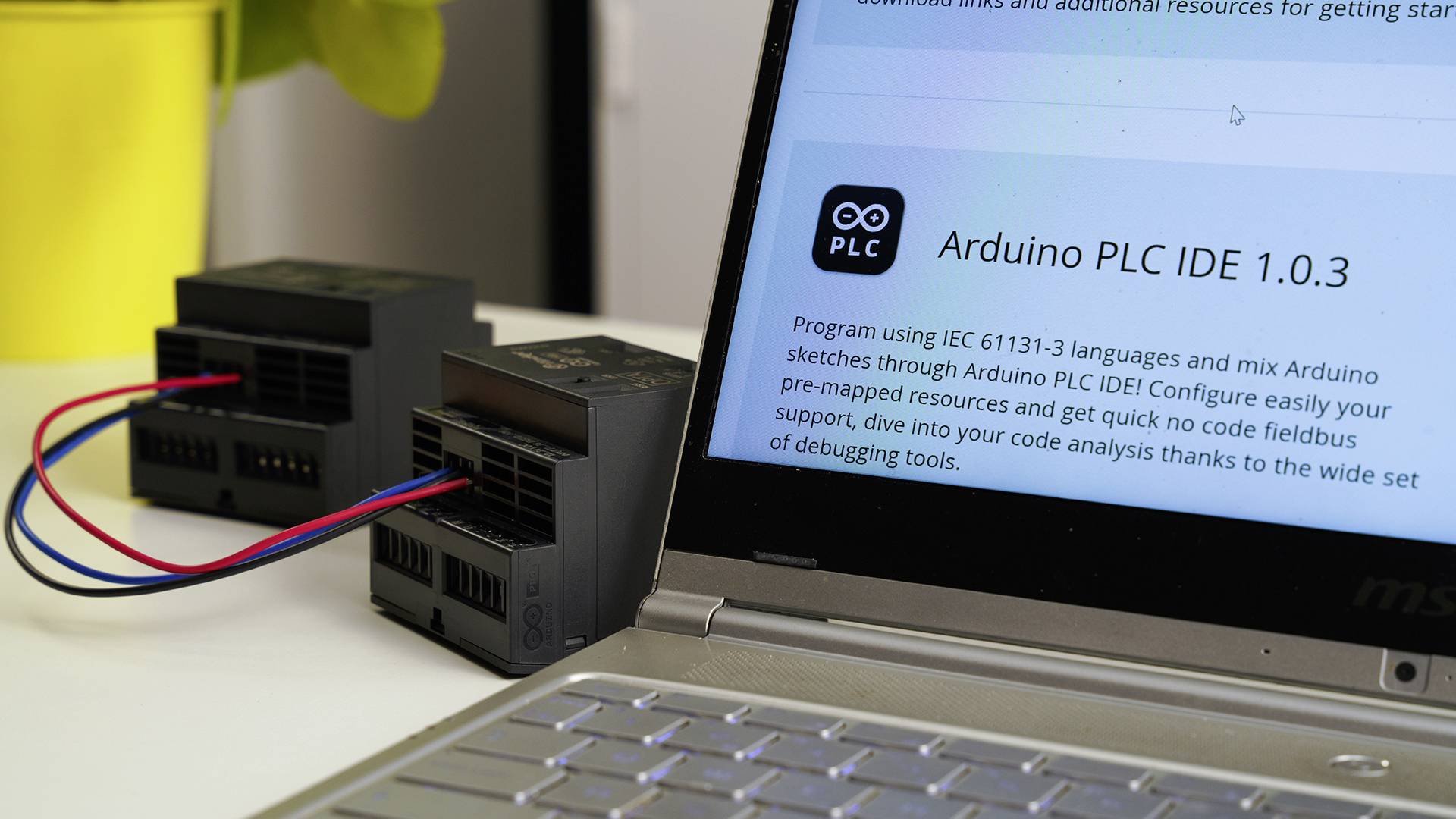 Laptop wyświetla stronę internetową z nagłówkiem 'Arduino PLC IDE 1.0.3'. Obok niego znajdują się dwa urządzenia elektroniczne podłączone przewodami.