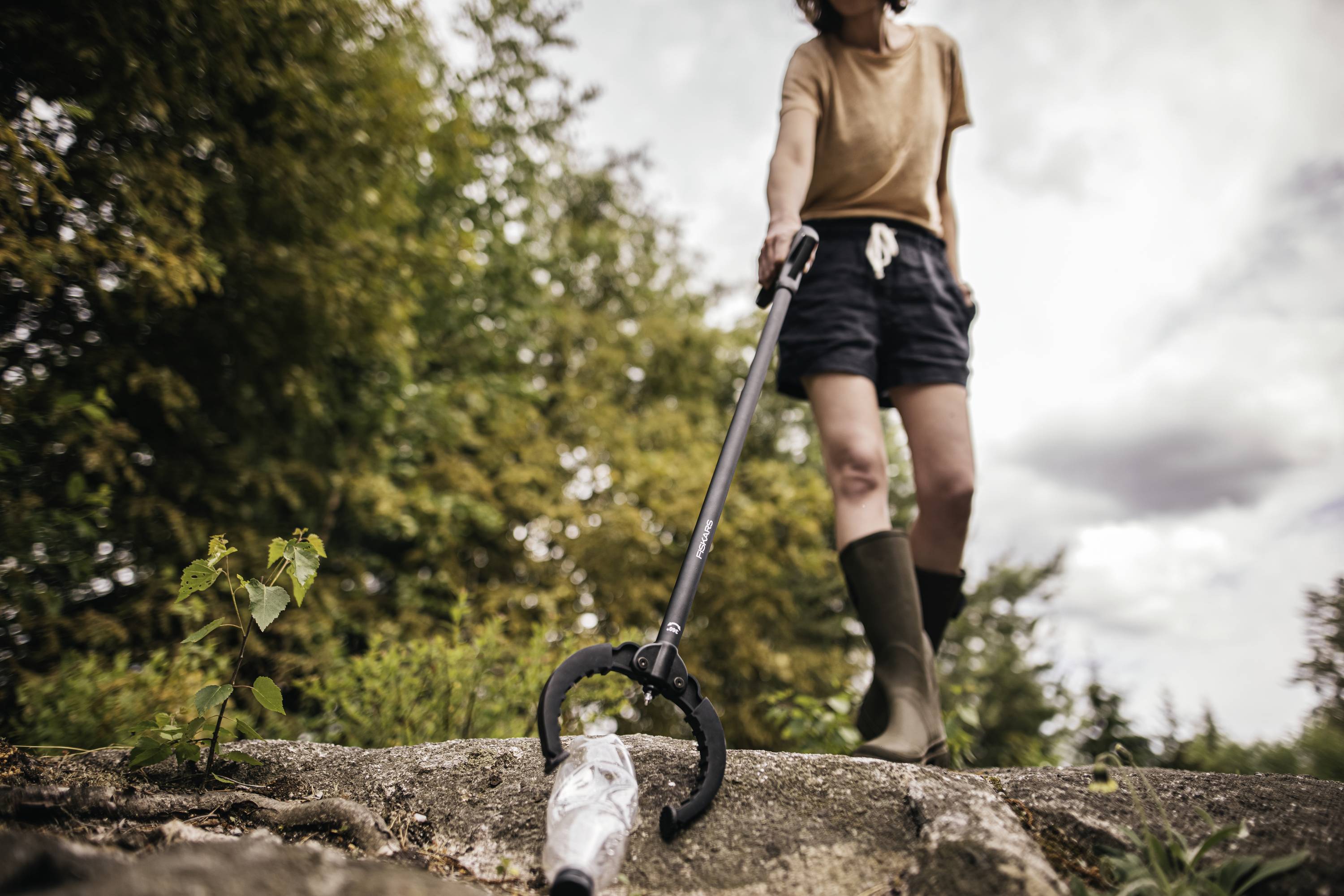 Osoba podnosi plastikową butelkę szczypcami w przyrodzie, ubrana w brązowy t-shirt, czarne szorty i gumowce.