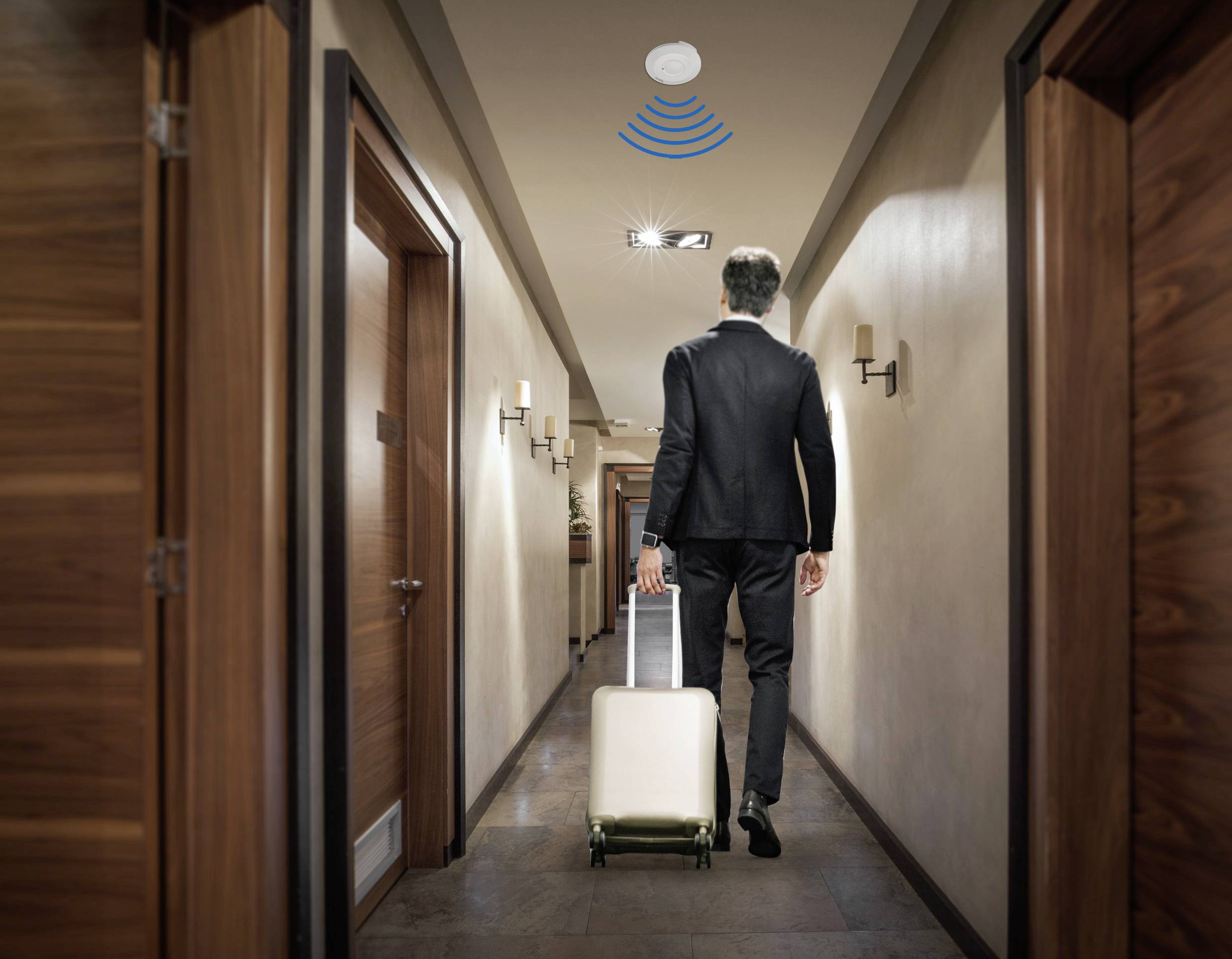 Mężczyzna w garniturze ciągnie walizkę przez korytarz hotelowy. Nad nim widać symbol Wi-Fi, wskazujący na inteligentną technologię.