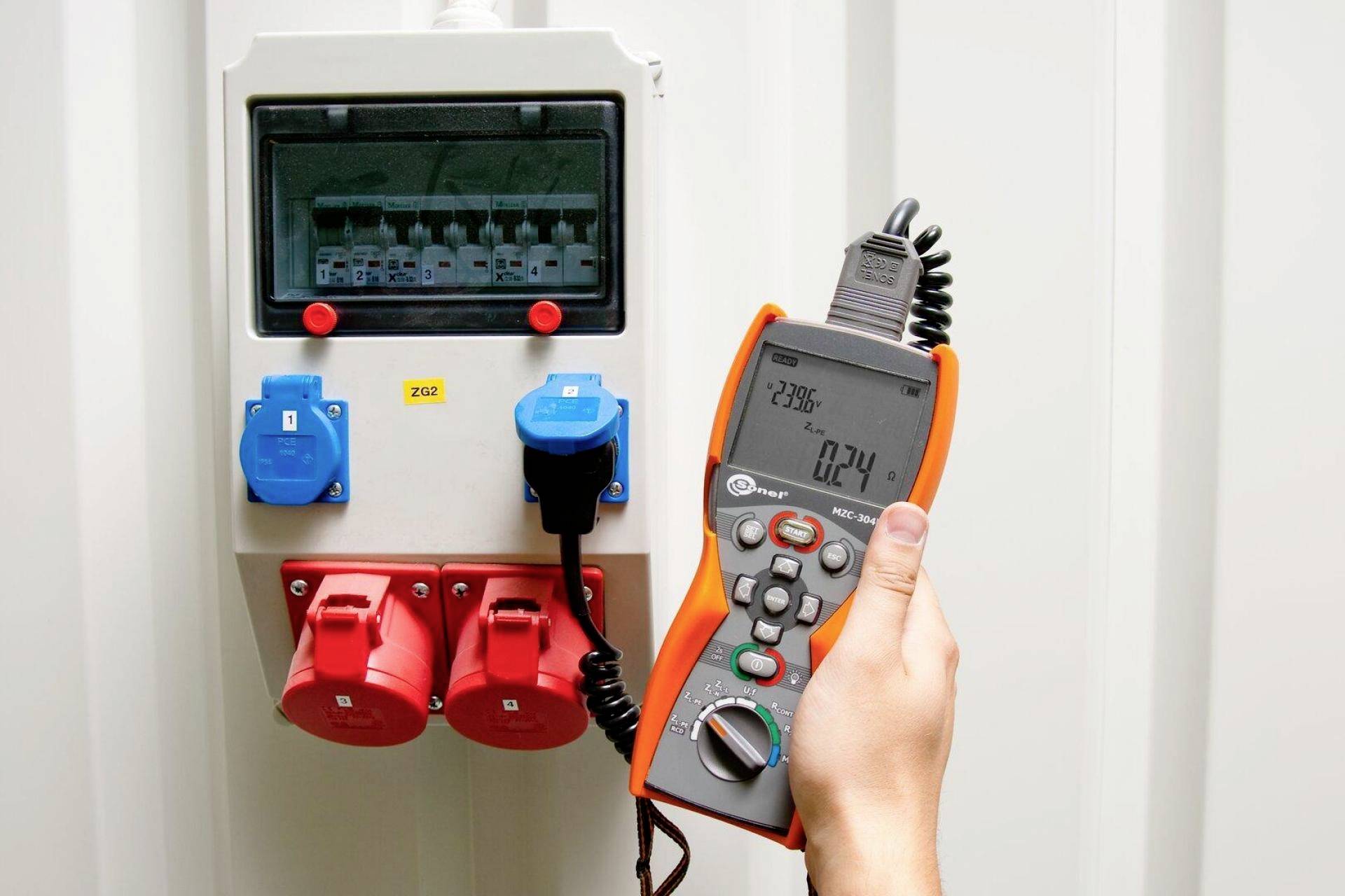 Osoba trzyma przyrząd pomiarowy w pobliżu szafy elektrycznej z czerwonymi i niebieskimi złączami. Urządzenie wyświetla wartości pomiarowe.