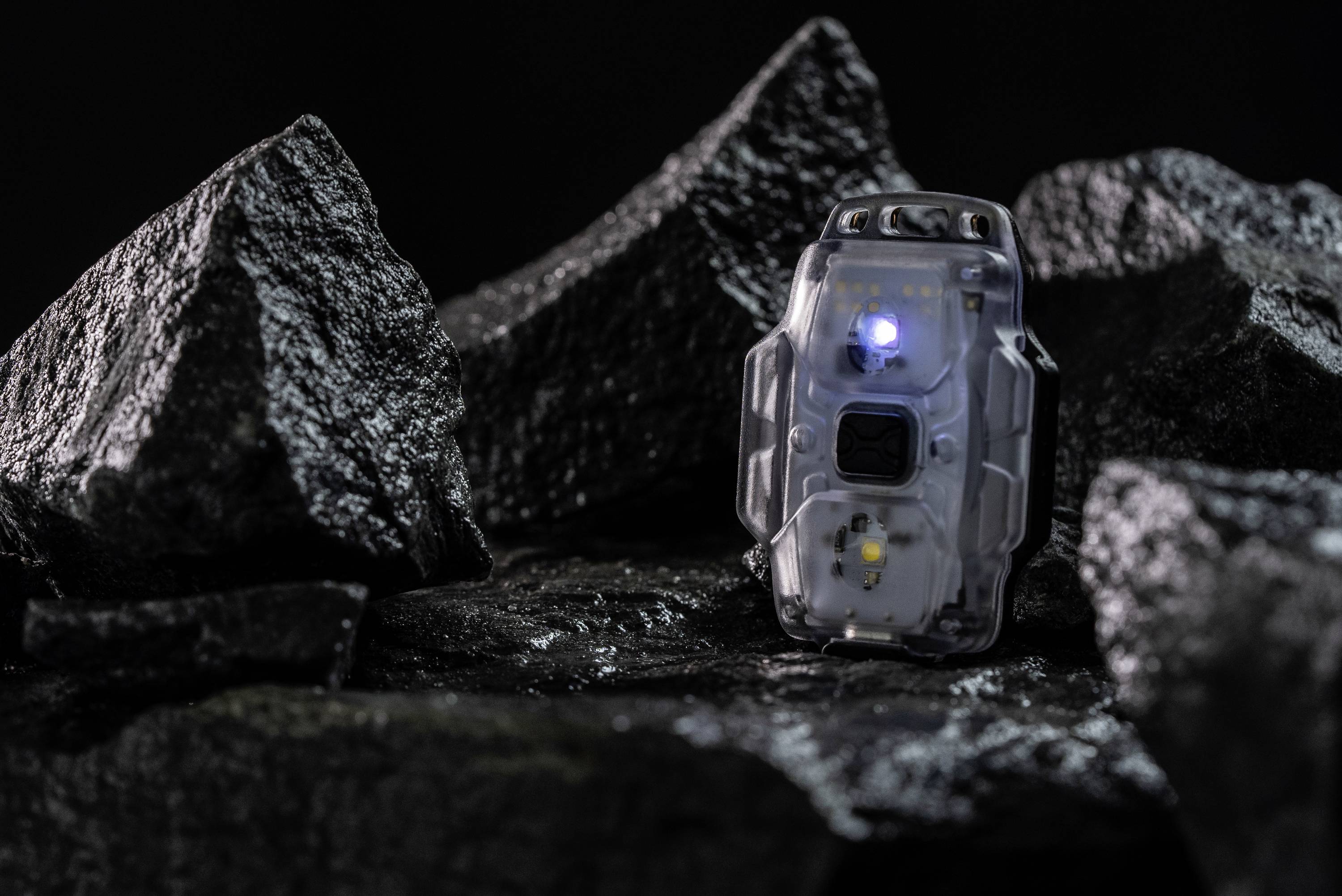 Mały, przezroczysty przyrząd z świecącą niebieską diodą LED leży na ciemnych, chropowatych kamieniach.