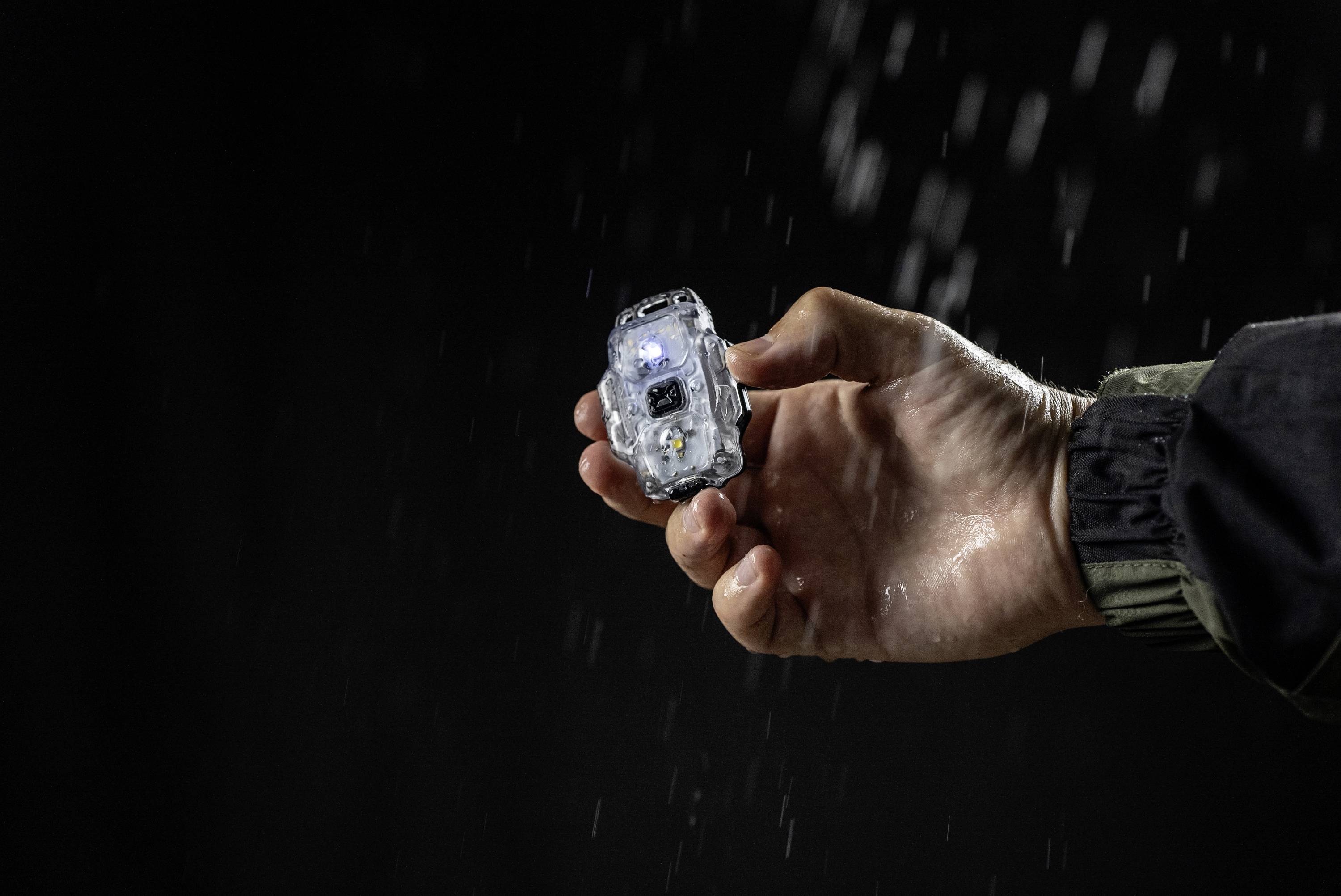 Dłoń trzyma małą, przezroczystą latarkę w deszczu, która świeci. Tło ciemne.