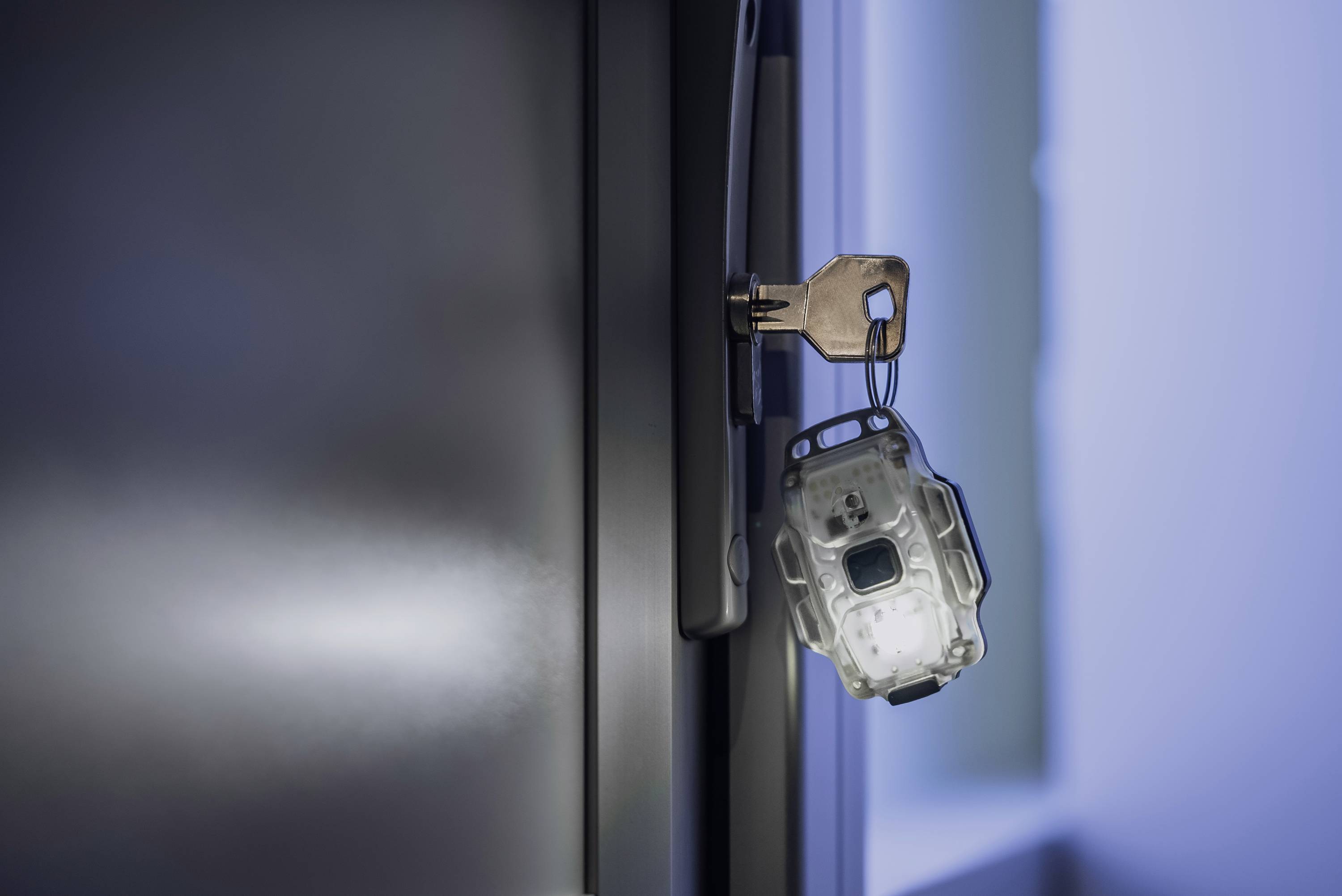Klucz tkwi w zamku drzwiowym, a przywiązany jest do niego przezroczysty brelok. Tło jest rozmyte i utrzymane w niebieskim odcieniu.