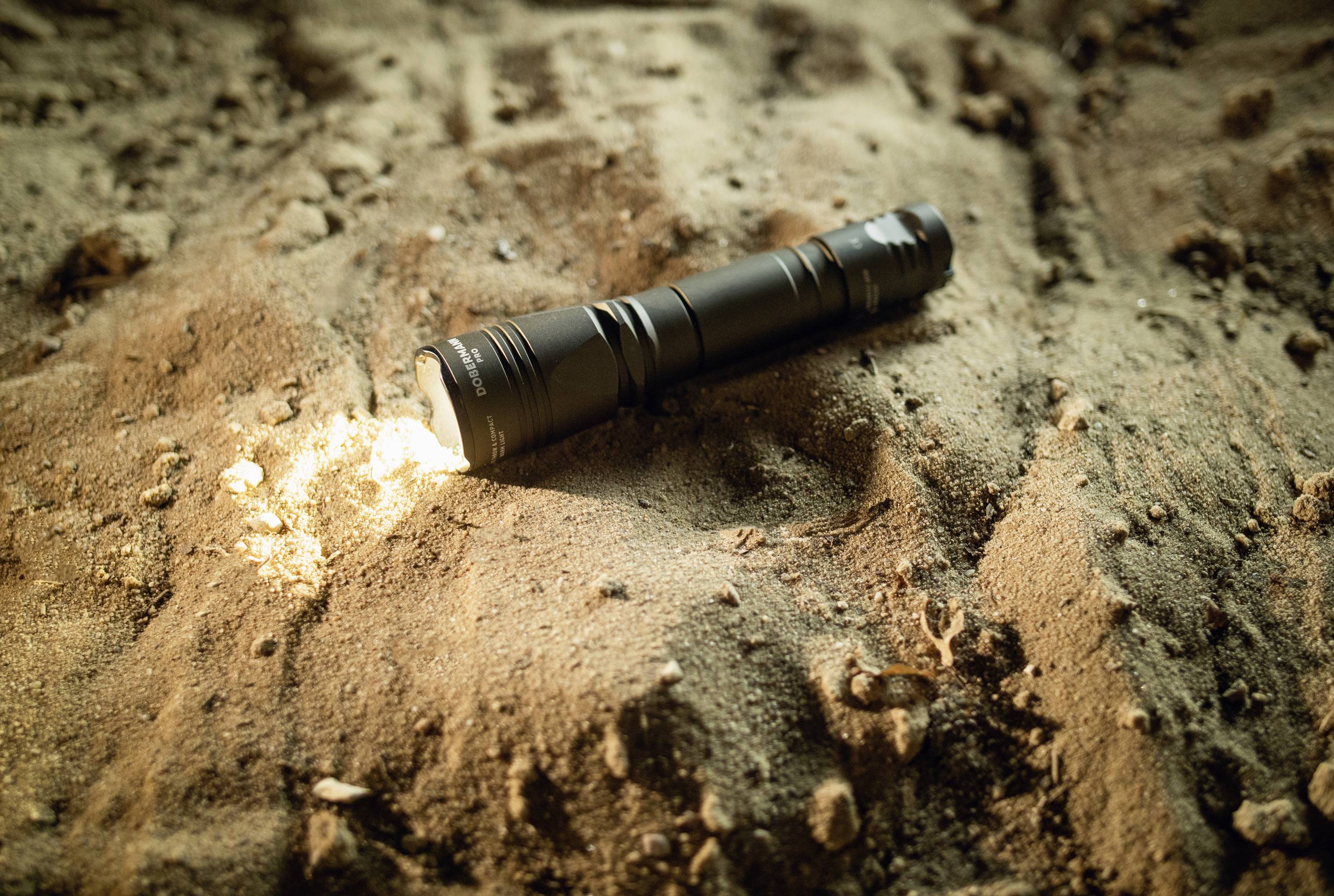 Latarka leży włączona na piaszczystym podłożu. Delikatnie oświetla piasek, pozostawiając mały krąg światła.