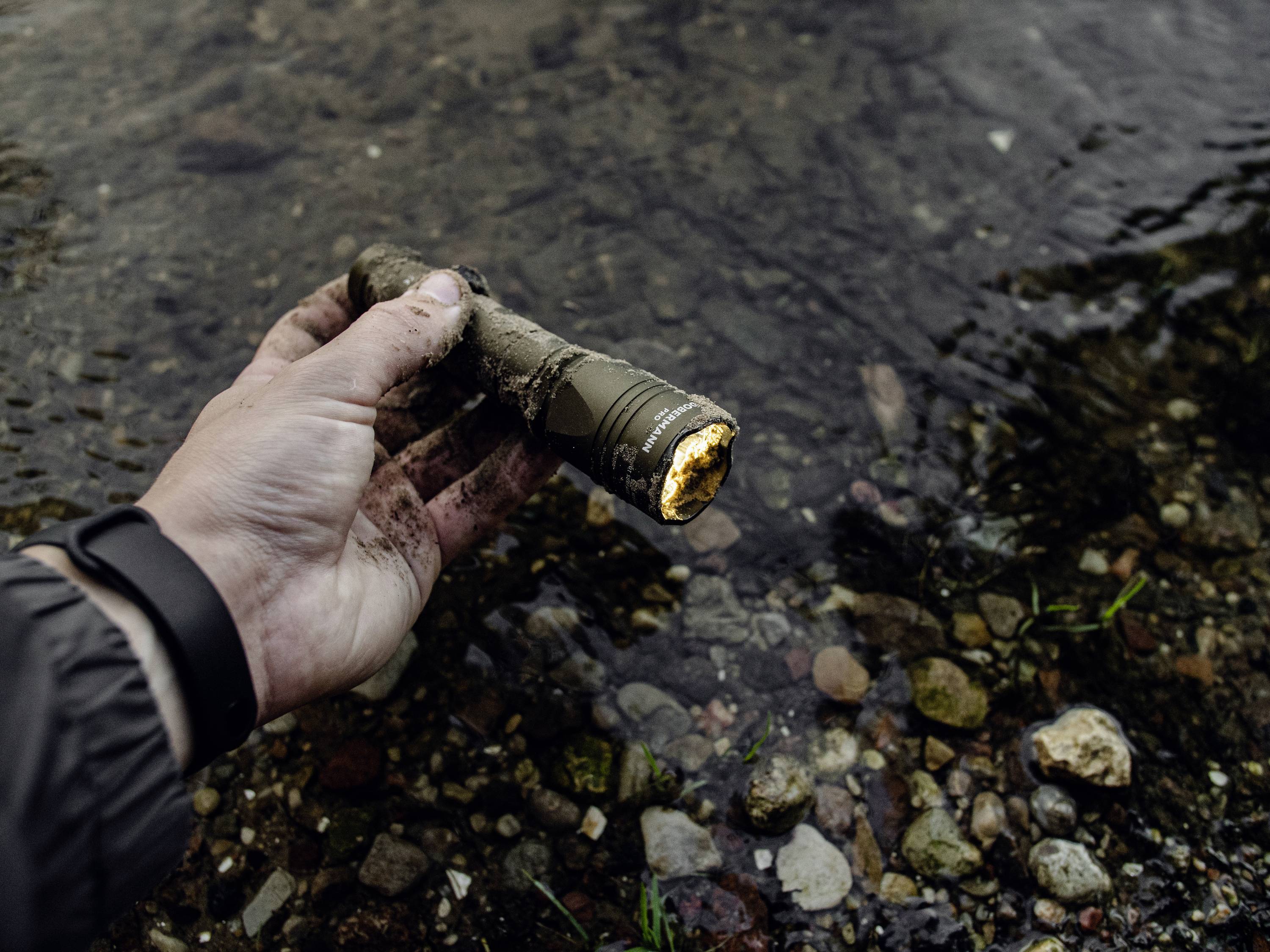 Dłoń trzyma brudną latarkę nad płaskim, kamienistym korytem rzeki. Latarka jest włączona.