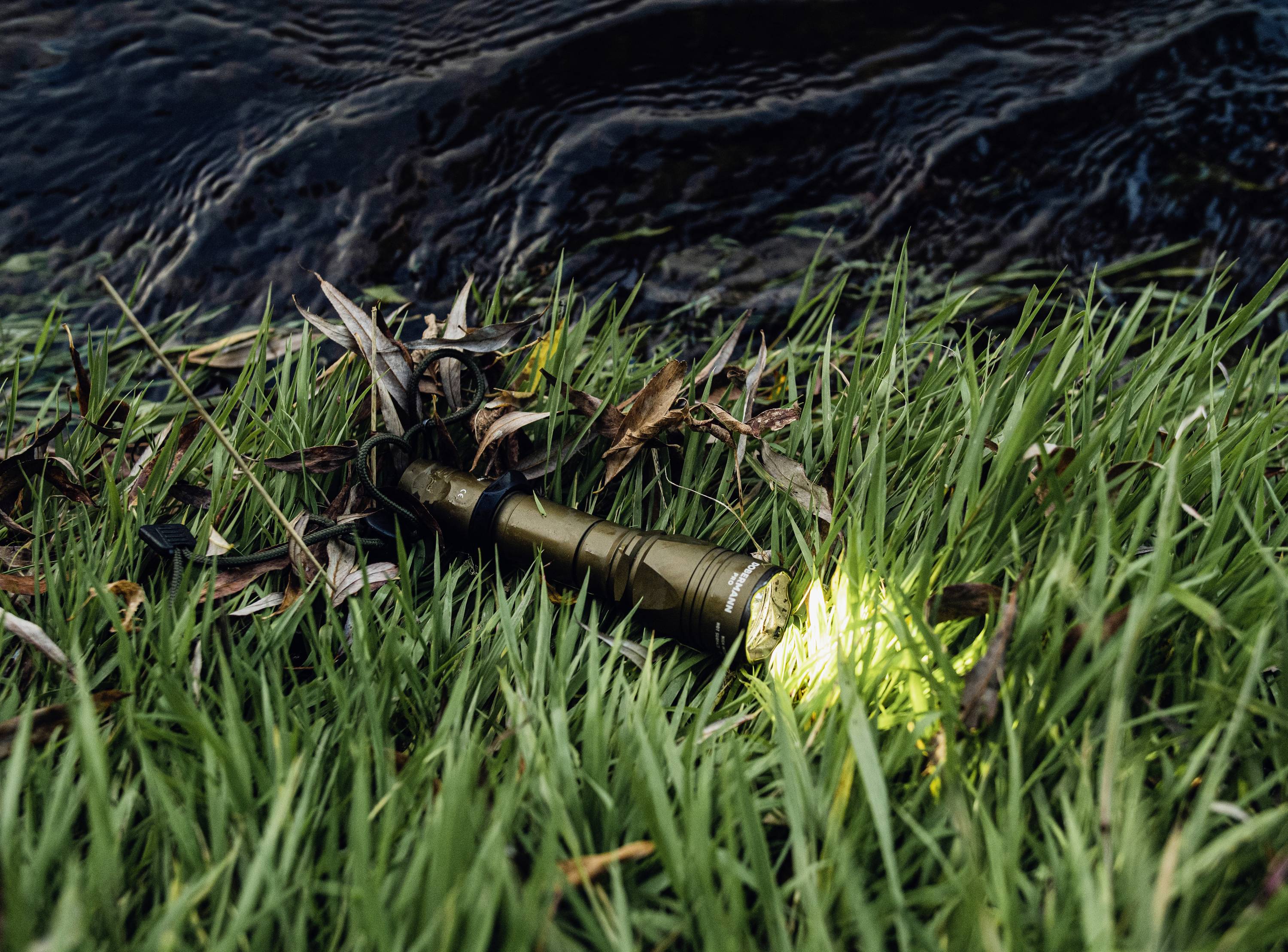 Latarka leży włączona na trawie obok zbiornika wodnego, a jej światło pada na okoliczne źdźbła i liście.