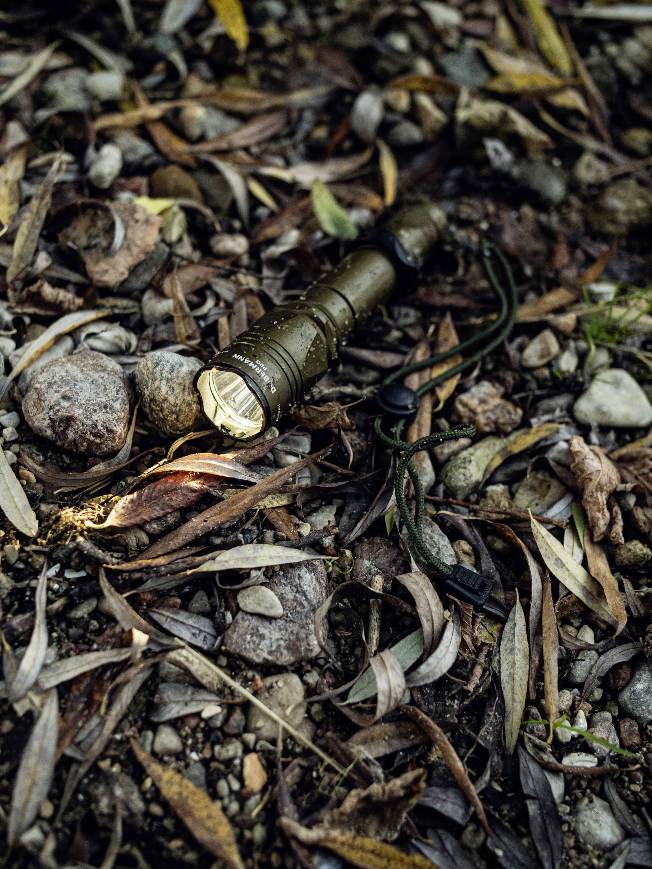 Latarka leży na leśnej ziemi, otoczona liśćmi i kamieniami, świecąc w prawą stronę.