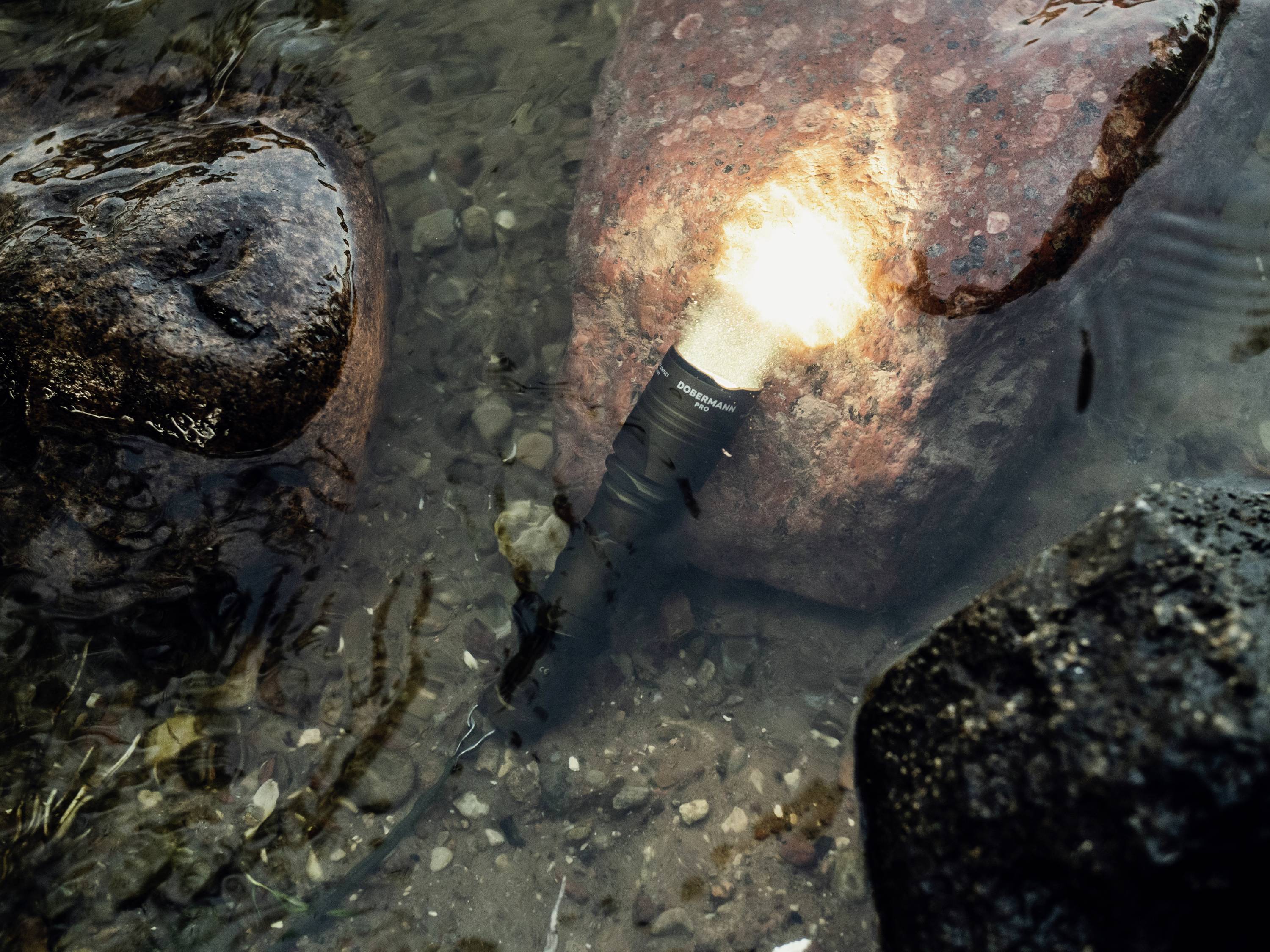 Świecąca latarka pod wodą, leżąca obok kilku kamieni w przejrzystej wodzie.