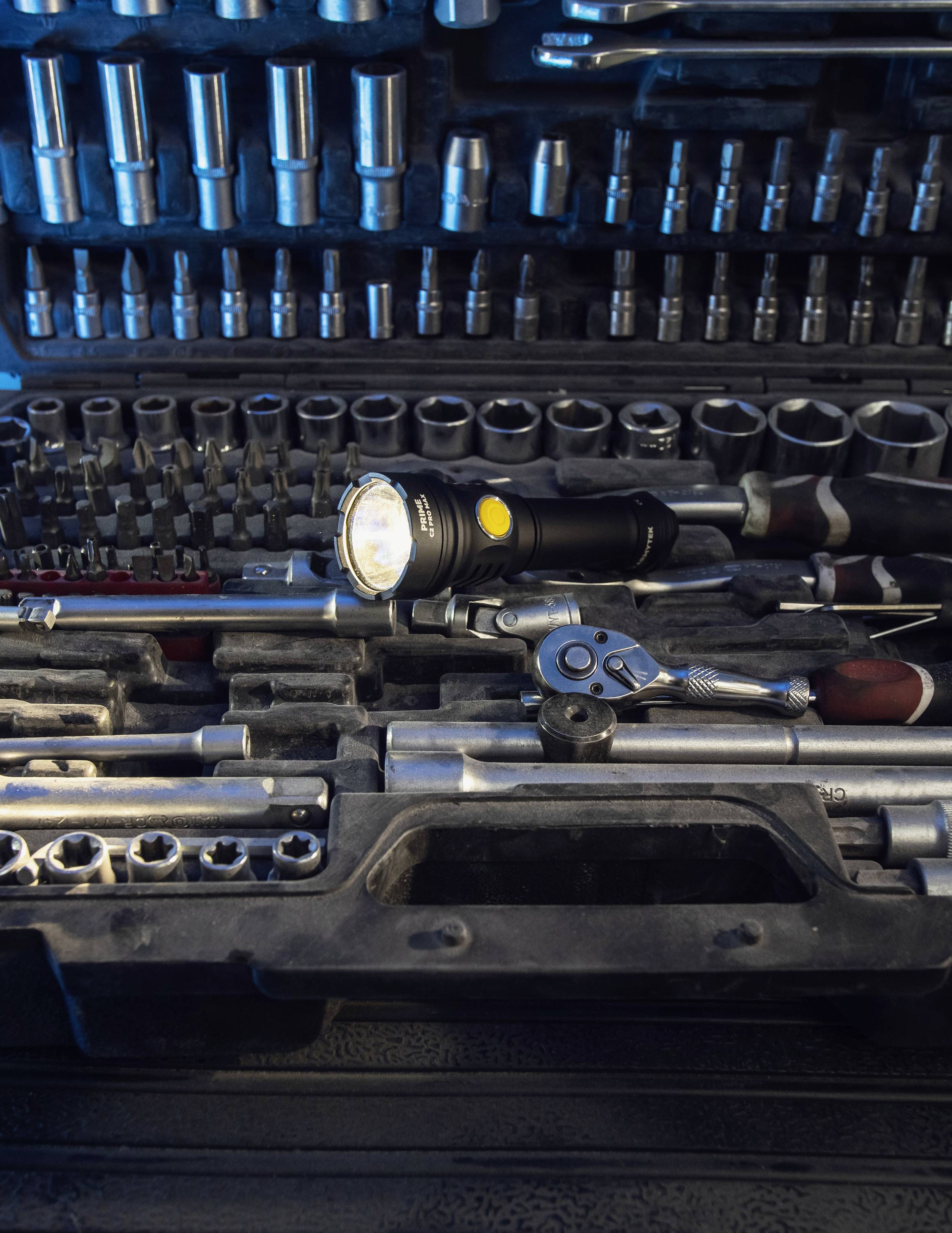 Czarna latarka świeci w skrzynce narzędziowej pełnej nakrętek i kluczy. Narzędzia są ułożone w sposób schludny i uporządkowany.