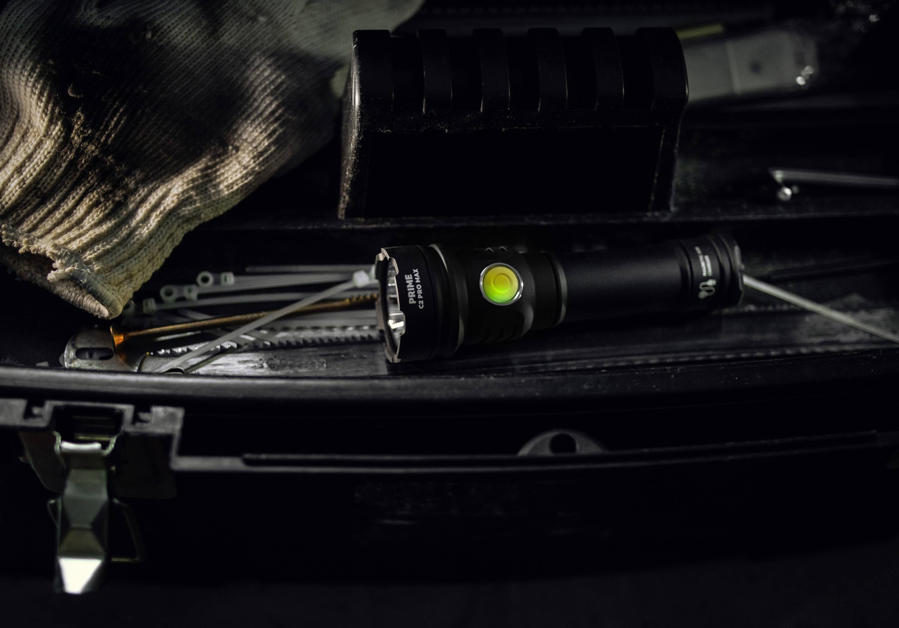 Czarna latarka leży w otwartej skrzynce narzędziowej, otoczona wkrętakami i szarą rękawicą roboczą.