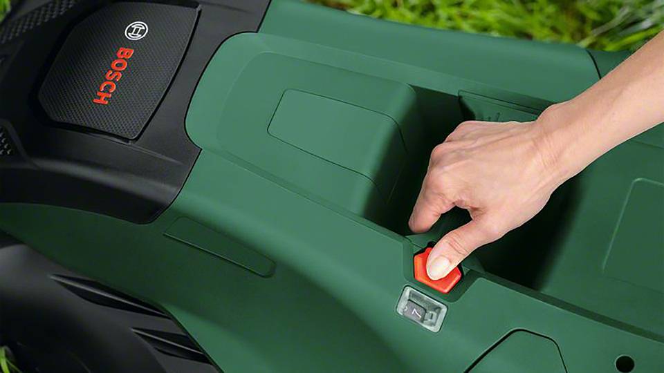 Osoba włącza zielony kosiarkę Bosch. Zbliżenie przedstawia rękę na czerwonym przycisku urządzenia, gotową do uruchomienia.