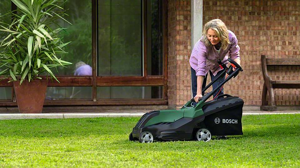 Kobieta kosi trawę boschowskim kosiarką w ogrodzie przed domem. W tle widoczne są okna i kamienny mur.
