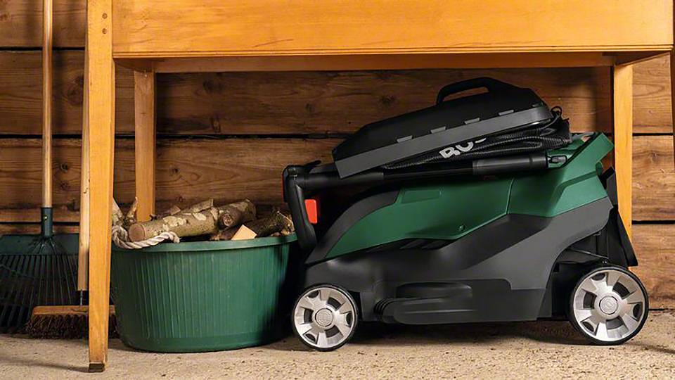 Kompaktowa, składana kosiarka w garażu obok kosza z drewnem opałowym i narzędziami ogrodowymi.