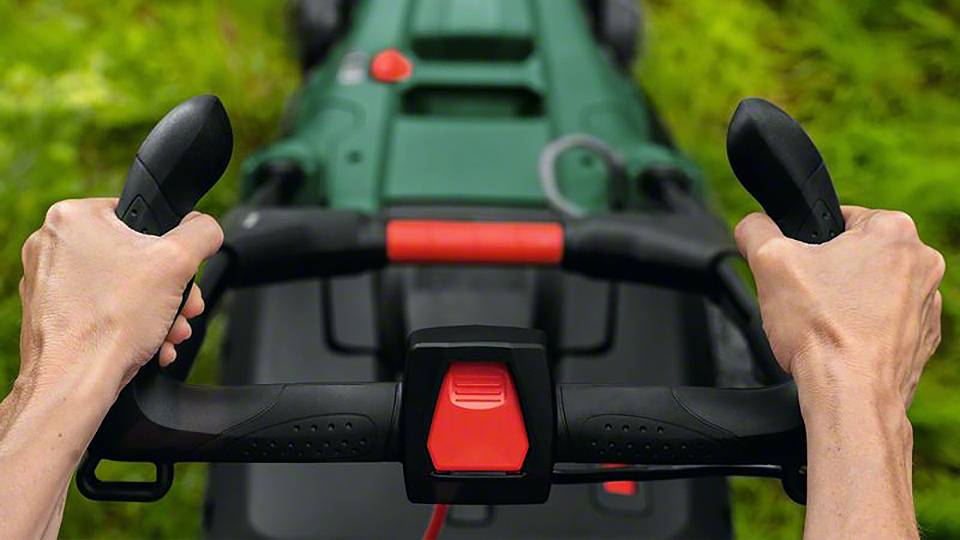 Dwie ręce trzymają kierownicę kosiarki, która stoi na zielonej trawie. Kosiarka posiada czerwone i czarne elementy sterujące.