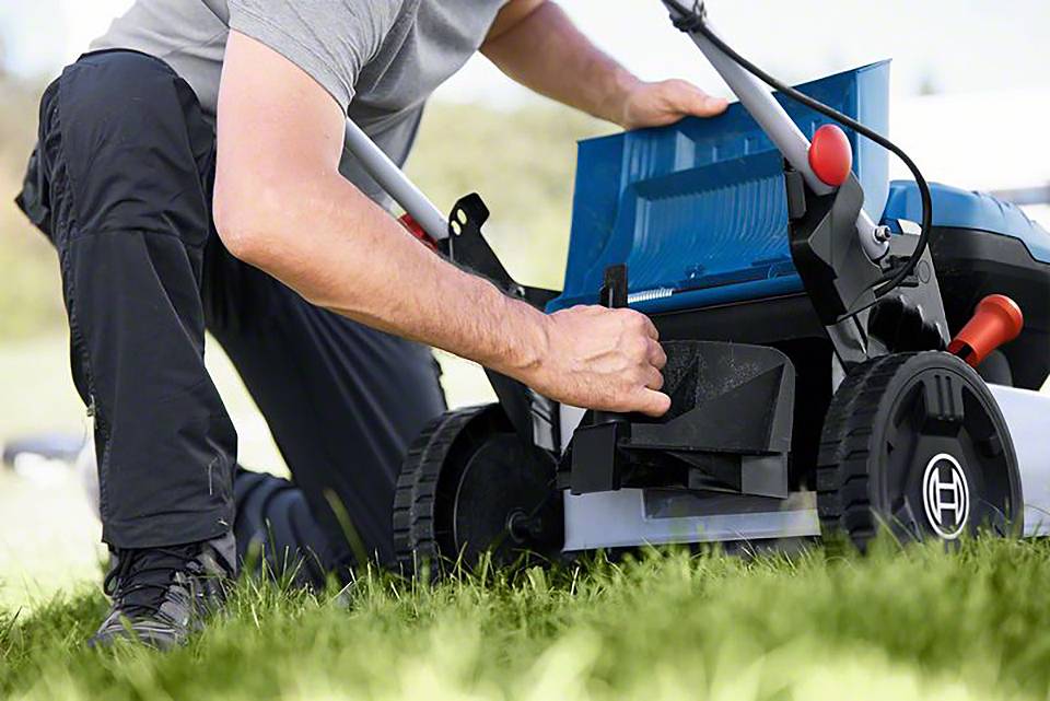 Osoba klęczy na trawniku i naprawia kosiarką. Fokus jest skierowany na dłoń, która sprawdza części urządzenia.