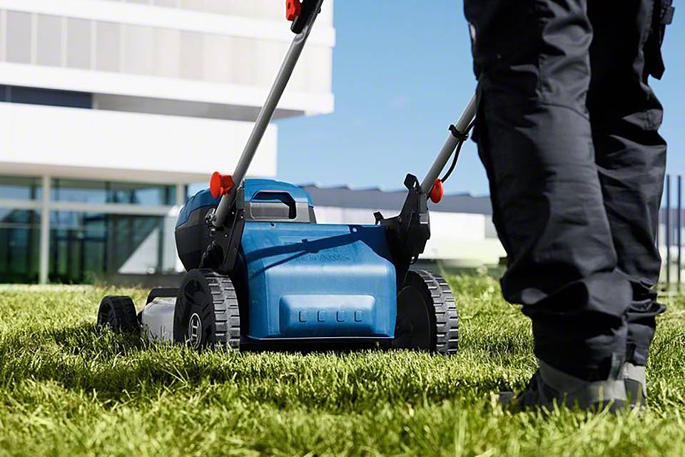 Mężczyzna kosi trawę niebieskim kosiarką w nowoczesnej przestrzeni na zewnątrz. W tle widoczne są budynki.
