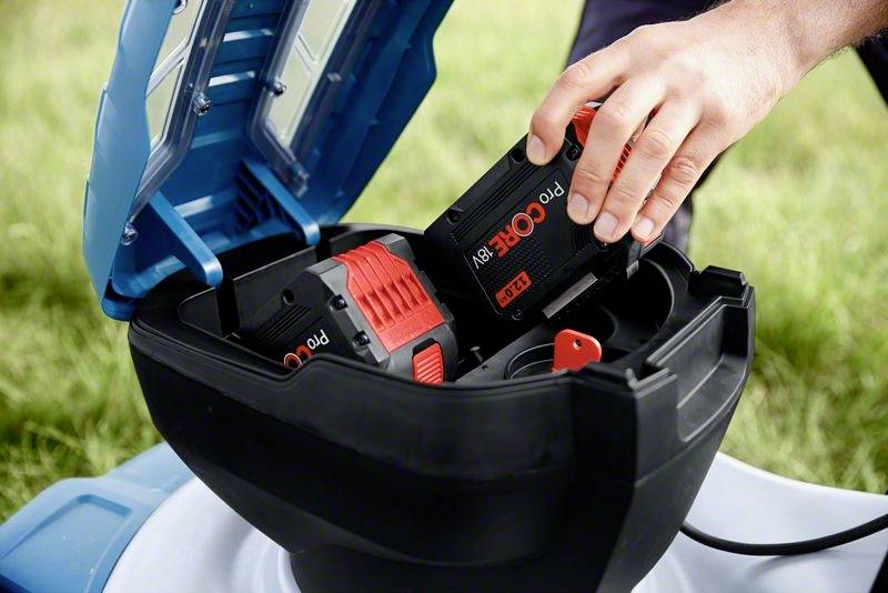 Osoba wkłada czarny i czerwony akumulator do kosiarki. Kosiarka ma niebieski pokrywa.