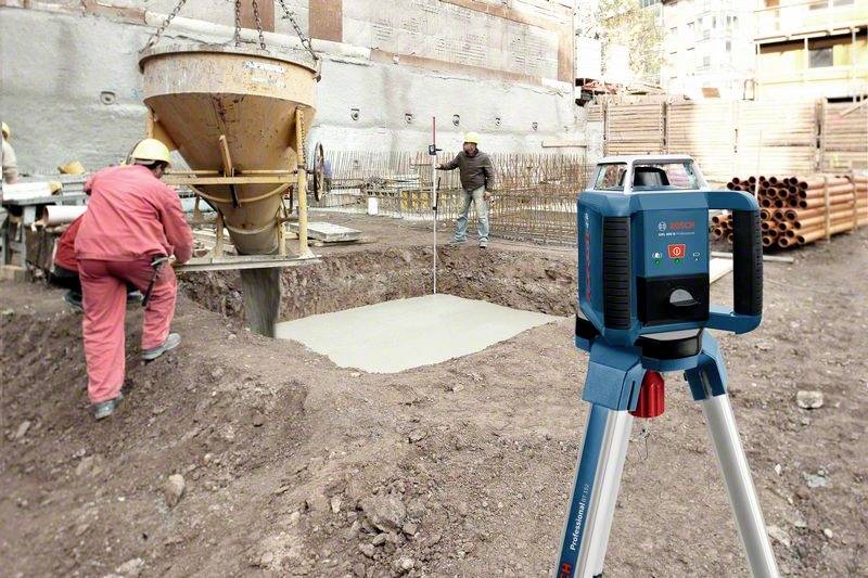 Plac budowy z robotnikami wlewającymi beton do wykopu. Na pierwszym planie stoi niwelator. W tle widoczne są budynki.