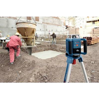 Plac budowy z robotnikami wlewającymi beton do wykopu. Na pierwszym planie stoi niwelator. W tle widoczne są budynki.