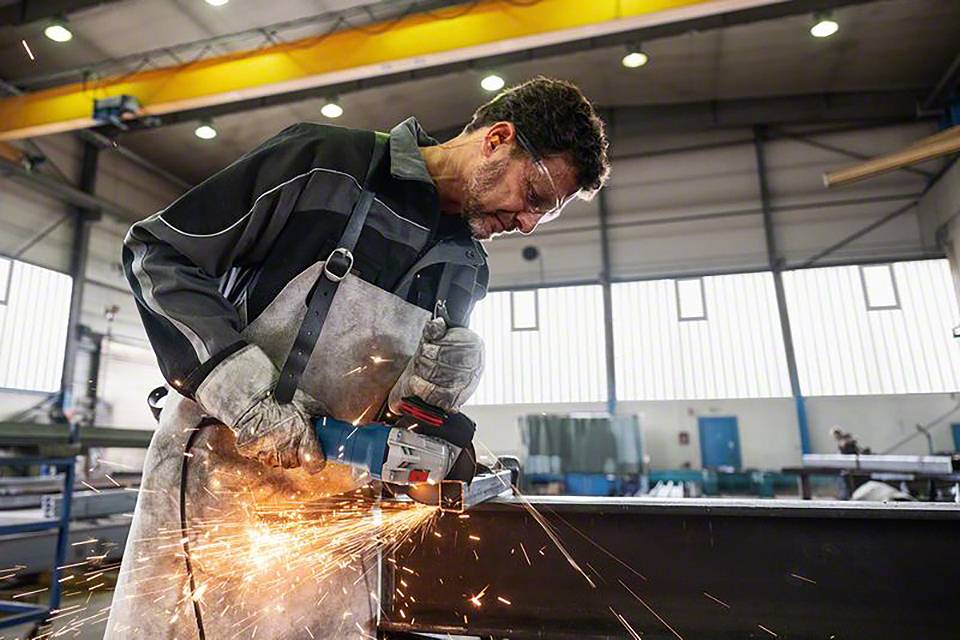 Mężczyzna w warsztacie ubrany w odzież ochronną używa elektronarzędzia do szlifowania, podczas gdy wokół niego lecą iskry.