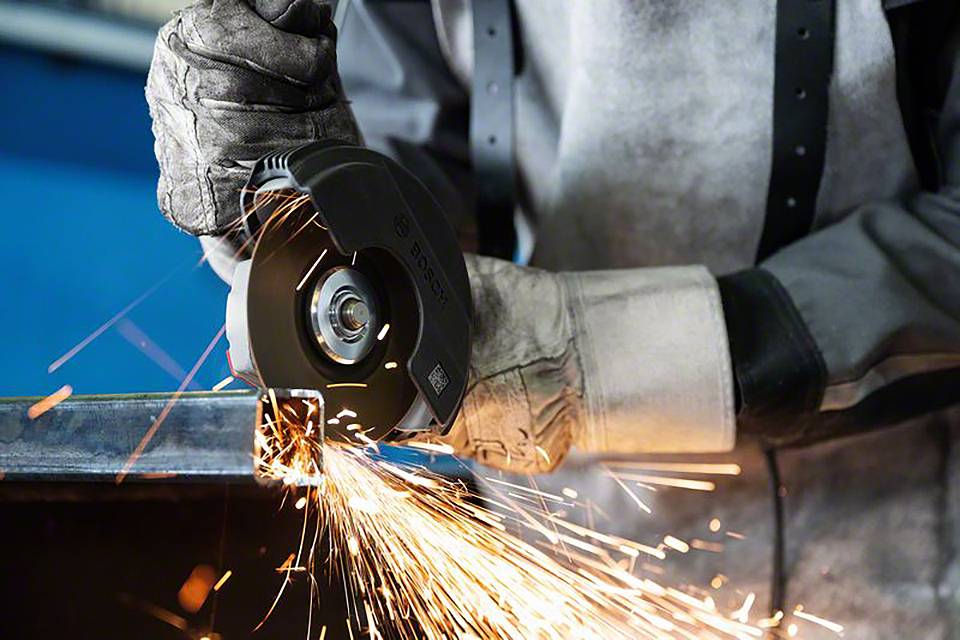 Osoba ucina metal szlifierką kątową, a wokół lecą iskry. Ma na sobie rękawice ochronne i odzież zabezpieczającą.