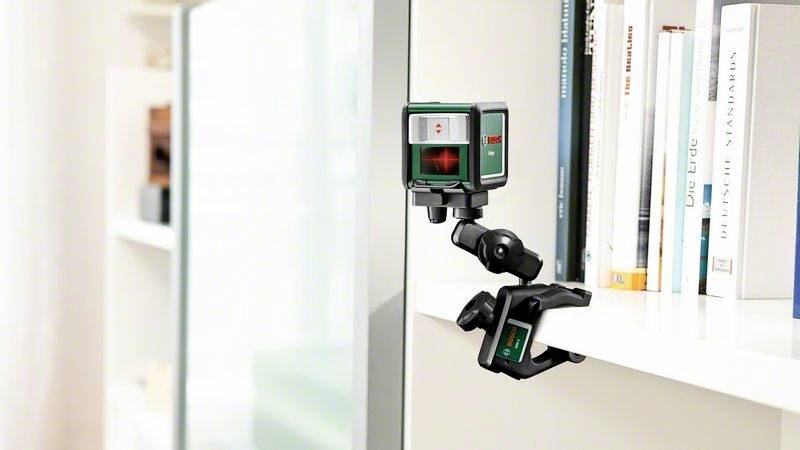 Laserowy miernik odległości jest zamocowany na regale i rzuca czerwoną wiązkę lasera na przeciwległy regał. W tle widoczne są książki.