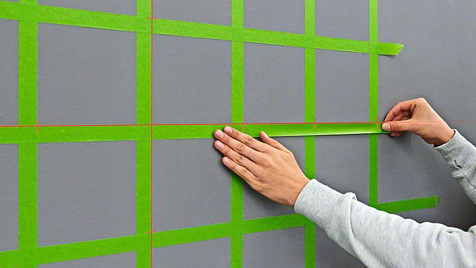 Osoba przykleja zielona taśmę klejącą w formie siatki na szarej ścianie, prawdopodobnie w ramach przygotowań do malowania lub dekoracji.