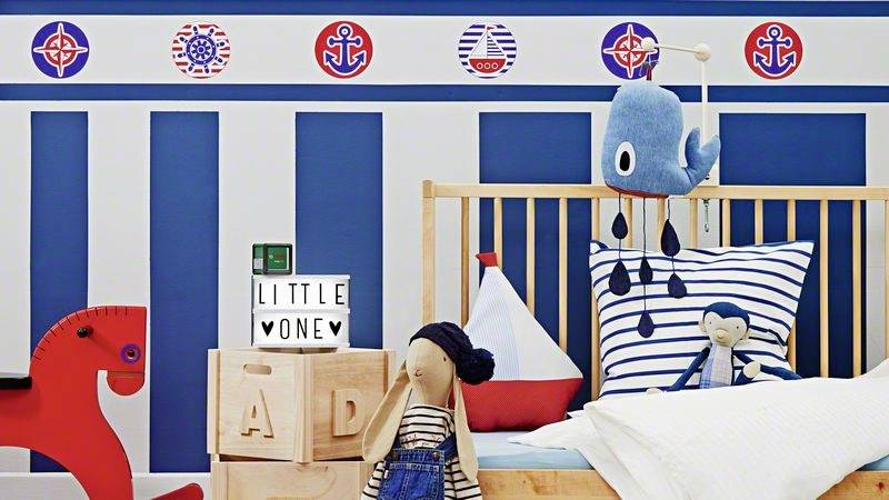 Pokój dziecięcy w stylu morskim: czerwona huśtawka, drewniane łóżeczko, poduszka z żaglówką, pluszowy wieloryb i królik, podświetlana skrzynka z napisem 'LITTLE ONE'.