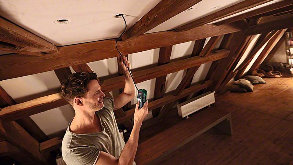 Mężczyzna na strychu instaluje lampę. Trzyma narzędzie i mocuje przewód do drewnianego belka nad sobą.