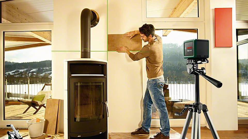 Mężczyzna mierzy ścianę obok pieca na drewno za pomocą urządzenia laserowego. Na zewnątrz widać zimowy krajobraz.