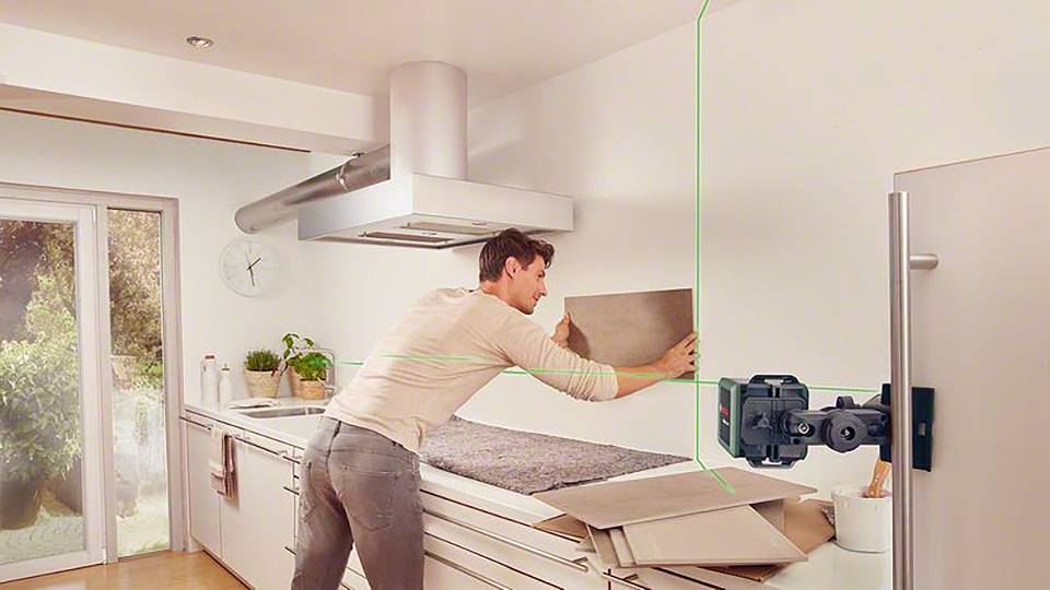 Mężczyzna wyrównuje kafel na ścianie kuchni za pomocą zielonego lasera poziomego. Kuchnia jest urządzona w nowoczesnym stylu.