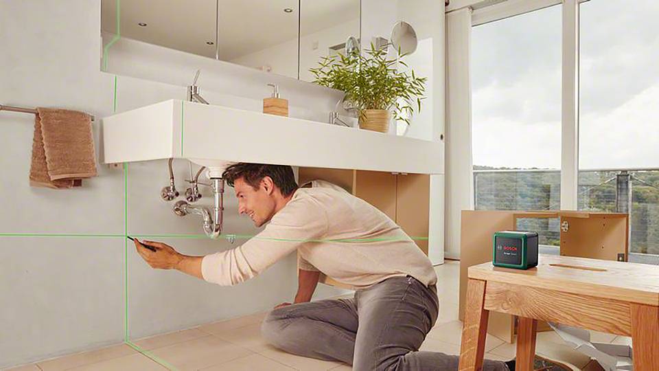 Mężczyzna reguluje miernik laserowy w łazience, stojąc przy umywalce. Zielony laser rzuca linie na ścianę.