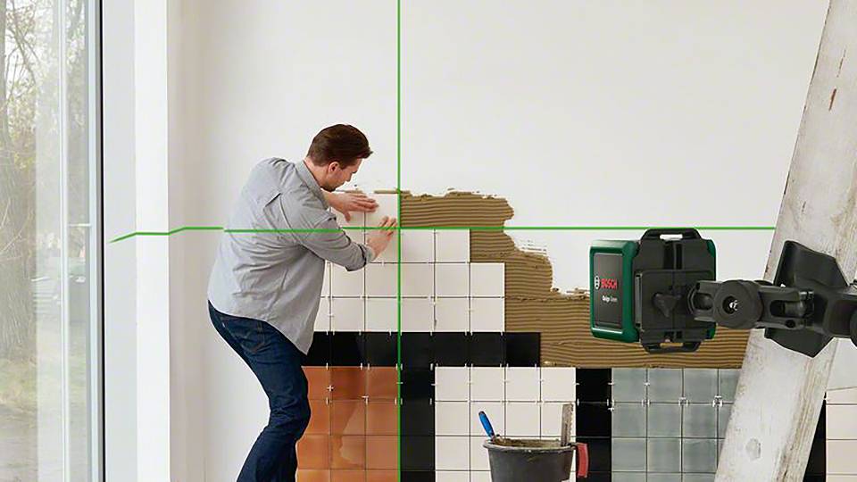 Mężczyzna nakłada płytki na ścianę, używając obok laserowego przyrządu do poziomowania. Po prawej stronie stoi drabina, a pod nią znajduje się wiadro zaprawy.