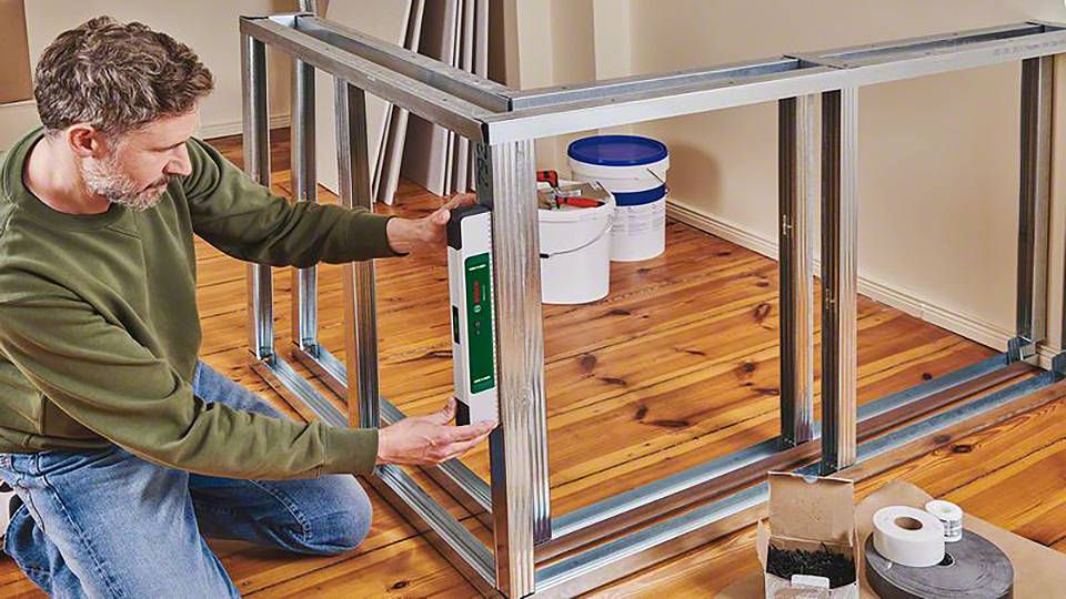 Osoba mierzy metalową ramę poziomicą w pomieszczeniu z drewnianą podłogą. W tle widoczne są wiadra i materiały.