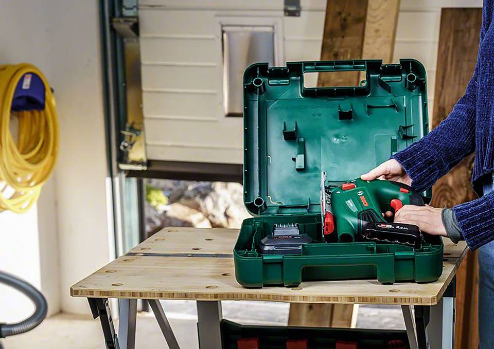 Osoba otwiera zielony kufer na narzędzia w warsztacie, w którym znajduje się akumulatorowy wkrętak i akcesoria, stojący na drewnianym stole.