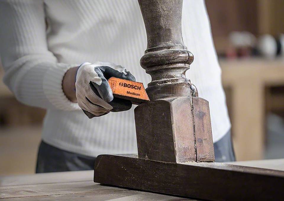 Jedna osoba wygładza drewniany słup za pomocą pomarańczowej gąbki ściernej Bosch. Ma na ręce rękawicę.