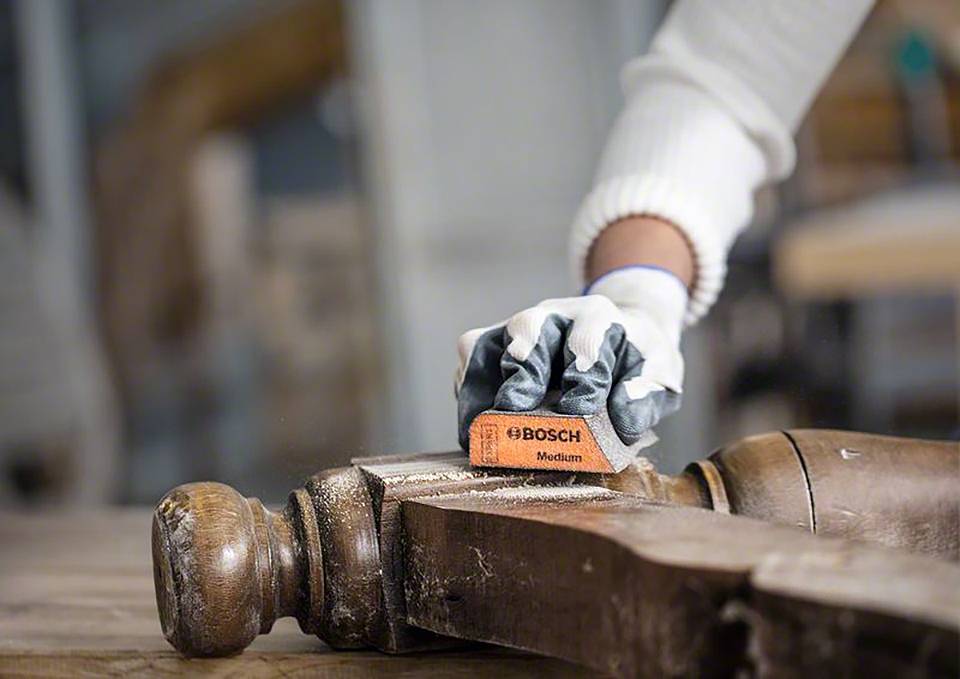 Osoba szlifuje kawałek drewna gąbką ścierną. Fokus jest skierowany na precyzyjną pracę przy detalu meblarskim.