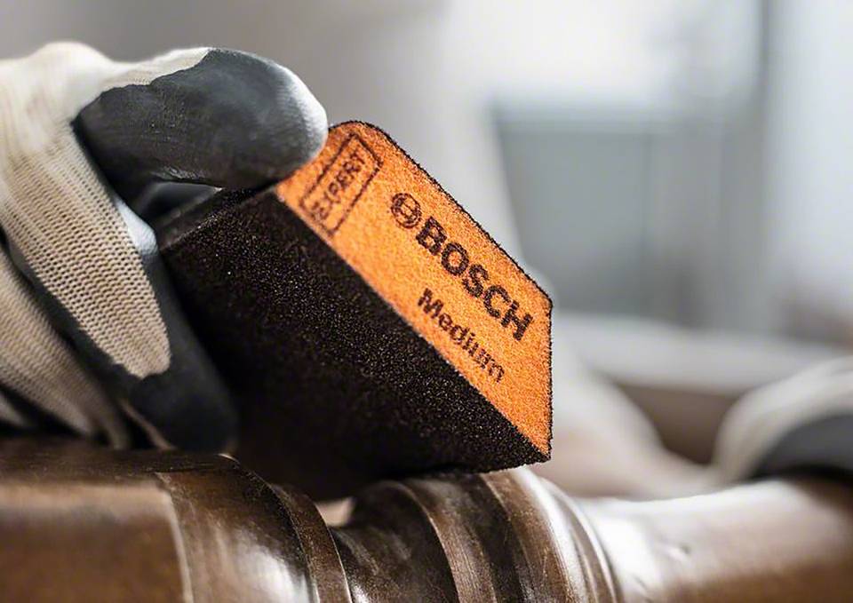 Ręka trzyma gąbkę ścierną Bosch z napisem 'Medium' i obrabia drewno. Rękawica służy do ochrony.