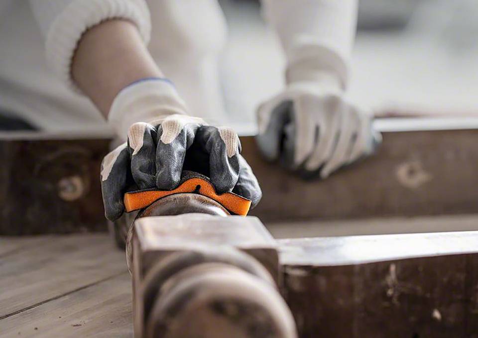 Osoba w rękawicach roboczych szlifuje kawałek drewna papierem ściernym na stole warsztatowym.
