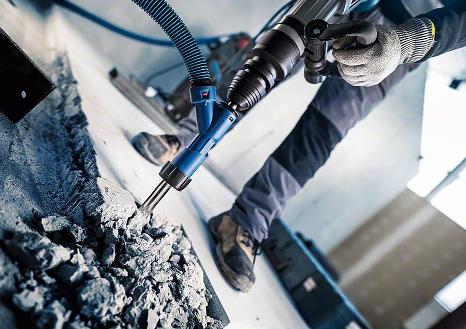 'Robotnik używa młotowiertarki do usuwania betonowej podłogi. Urządzenie przeciwpyłowe na wiertarce zmniejsza wytwarzanie się pyłu.'