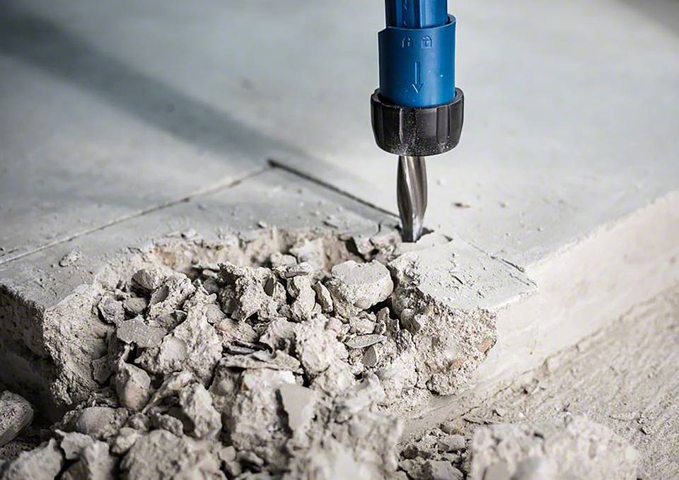 Wiertło obrabia beton, wokół miejsca wiercenia widoczne są kawałki betonu i pył.