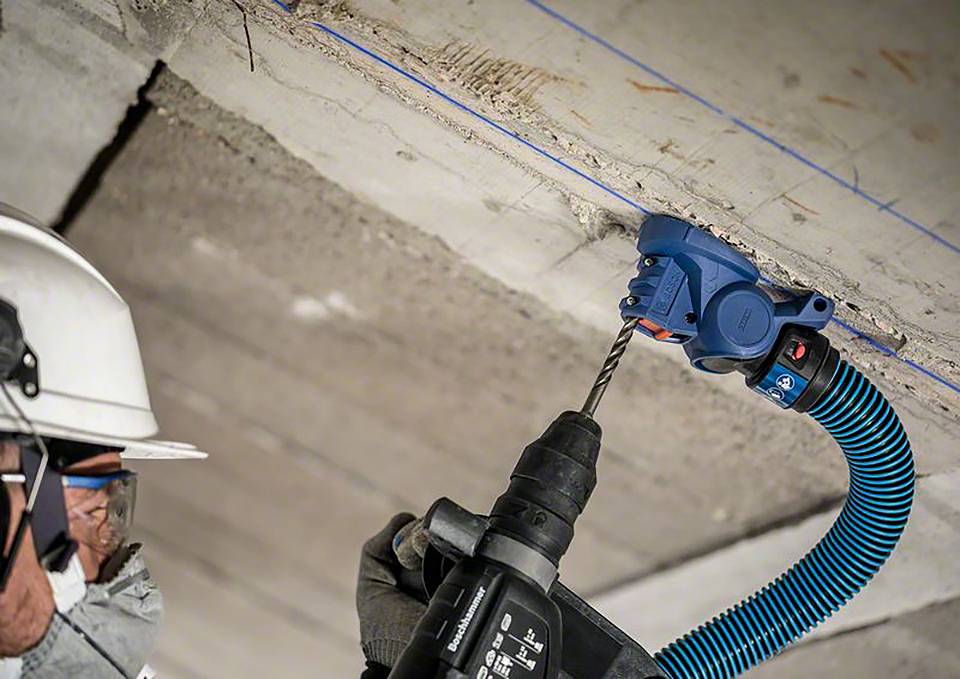 Robotnik budowlany w odzieży ochronnej wierca otwór w stropie betonowym za pomocą elektronarzędzia, podczas gdy zamontowany jest odkurzacz przemysłowy.