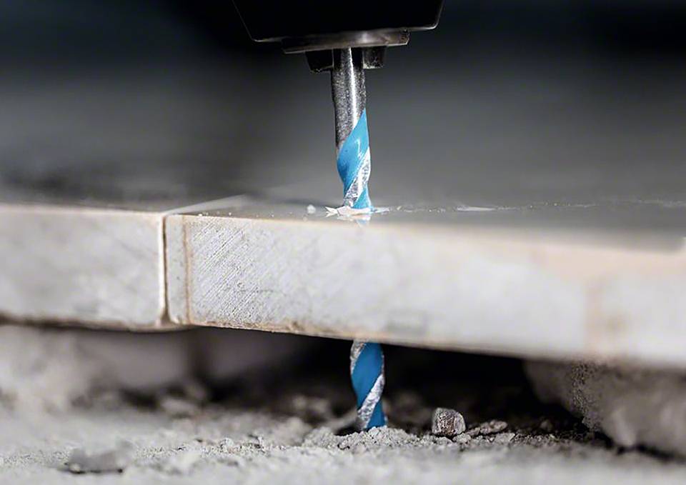 Wiertło przebija płytkę, otoczone pyłem i gruzem. Zbliżenie ukazuje niebieski i metaliczny wiertarz w akcji.