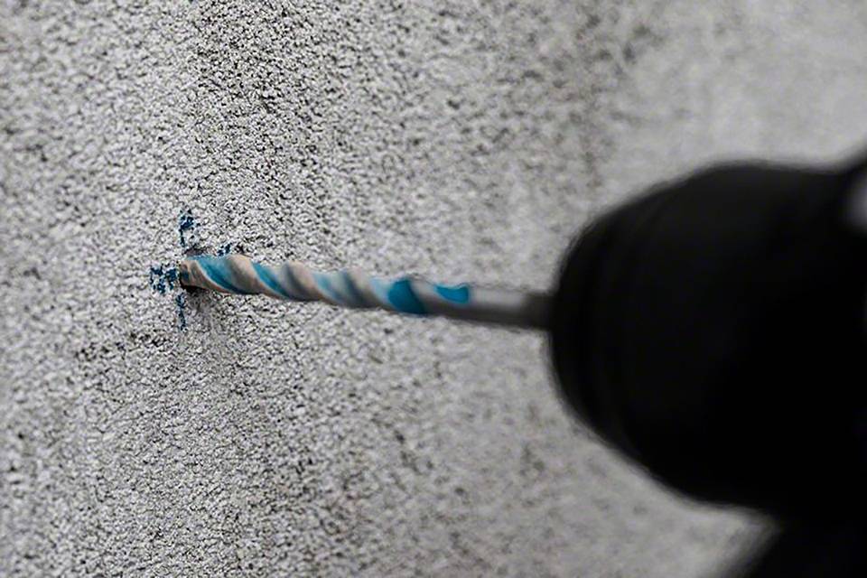 Wiertło przewierca szorstką, szarą ścianę, tworząc drobny pył. Główna czynność: wiercenie w twardej powierzchni.