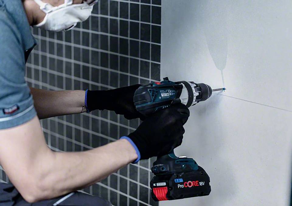 Osoba wierci dziurkę w ściance kaflowej za pomocą elektrycznej wiertarki. Ma na sobie rękawice i maskę.