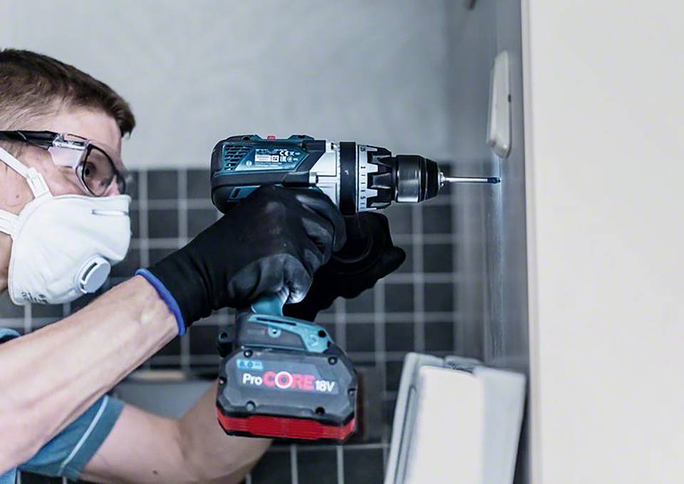 Mężczyzna w okularach ochronnych i masce używa elektrycznej wiertarki, aby wywiercić otwór w wykafelkowanej ścianie.