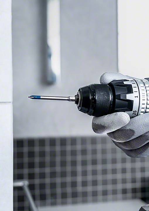 Jedna ręka trzyma wiertarkę, która jest gotowa, aby wywiercić otwór w wykafelkowanej ścianie. W tle nieostro widać łazienkę.