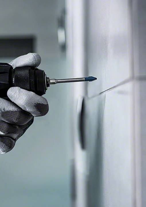 Dłoń w rękawicy trzyma akumulatorową wkrętarkę przyciśniętą do wykafelkowanej ściany, aby wywiercić otwór.