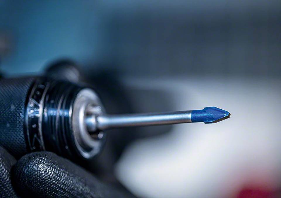 Zbliżenie maszyny wiertniczej z niebieskim wiertłem, trzymanej przez osobę w czarnych rękawicach roboczych.