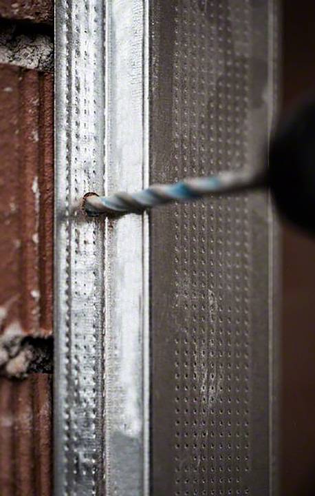 Wiertarka wierci otwór w belce metalowej przy ścianie z cegły. Belka metalowa jest częścią konstrukcji budowlanej.